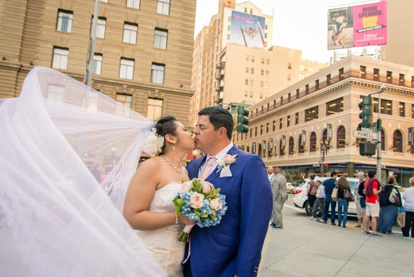 Bride and groom kissing on sidewalk, her flowing veil blowing in the breeze in downtown San Francisco.