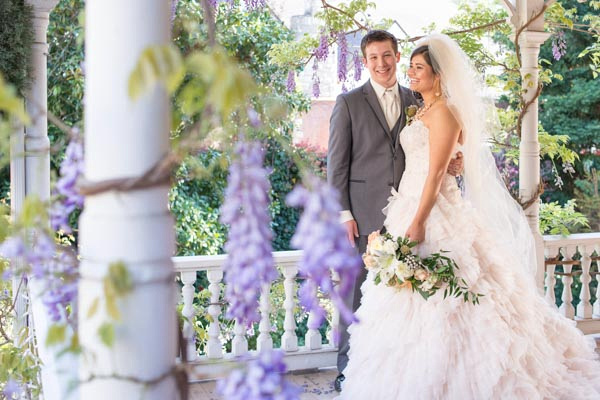 Young bride and groom pose on white Victorian balcony surrounded by vines with purple blossoms at The Seasons Bed & Breakfast in Placerville.